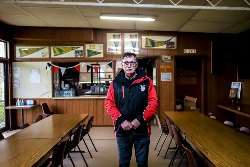 Older man with grey moustache and glasses stands in brown wooden room with memorabilia behind him