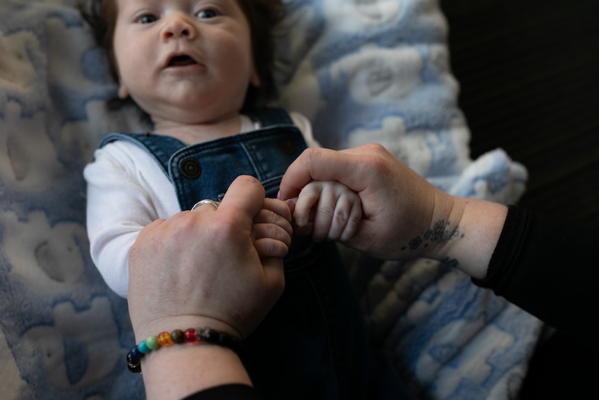 A baby lying down looks up at the camera, his mouth open. His mother's hands are holding his.