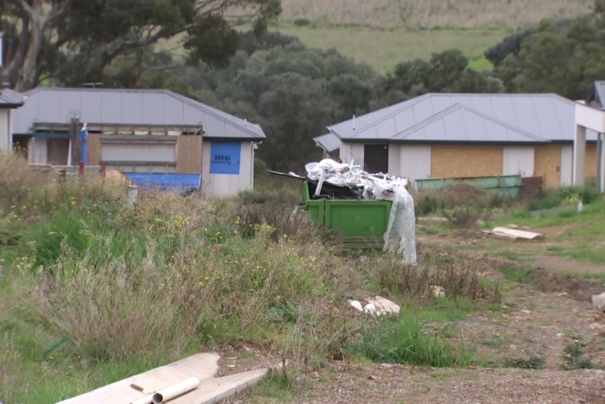 Houses under construction with long grass and a skip bin filled with rubbish