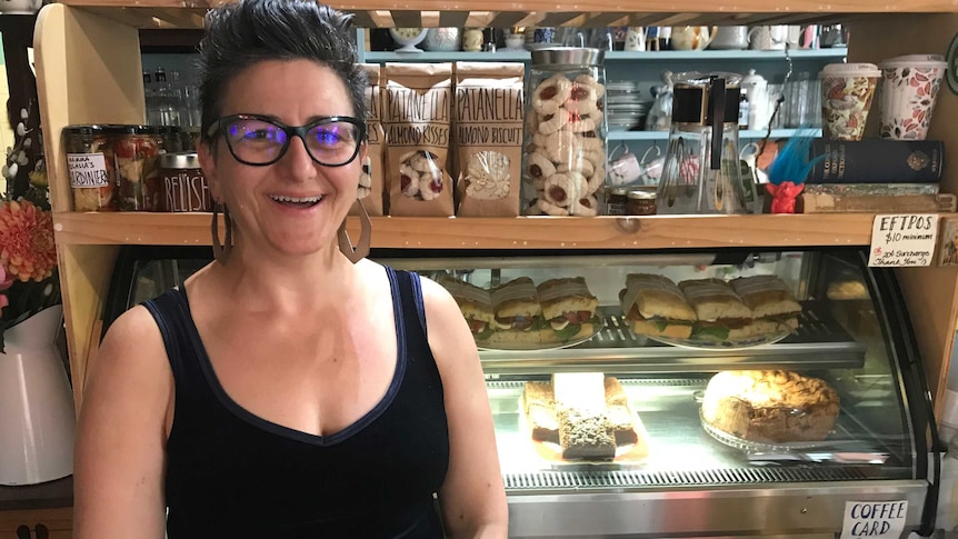 woman smiles brightly in front of cafe display fridge of cakes and savouries