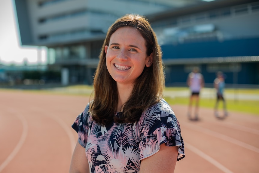 A young woman with long brown hair smiling at the camera while at an athletics track