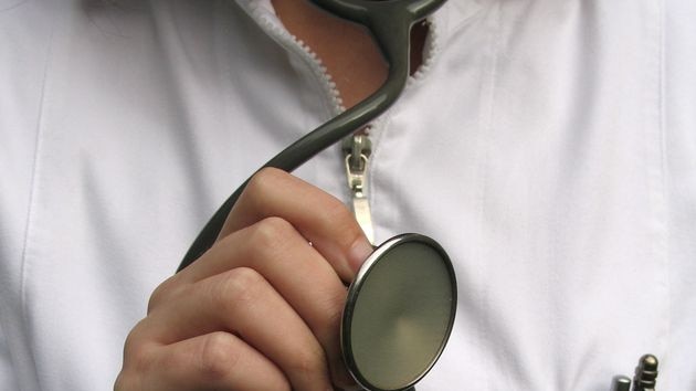 An unidentified female doctor in a white coat holds a stethoscope towards the camera.