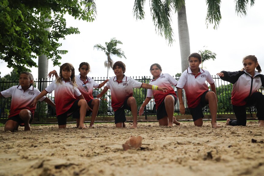 Children in red and white shirts and black shorts kneel on sand in a choreographed dance routine