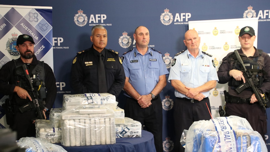 A line of police, some armed, standing behind plastic wrapped packaged piles of cocaine.