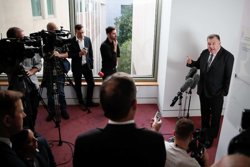 A man talks behind microphones surrounded by journalists and camera