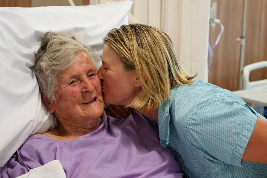 A smiling Barbara Marincic looks up from her hospital bed as her daughter kisses her on the cheek