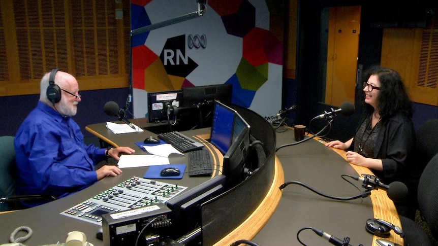 Adams with headphones on and Kats-Chernin sitting opposite each other in radio studio in front of microphones.