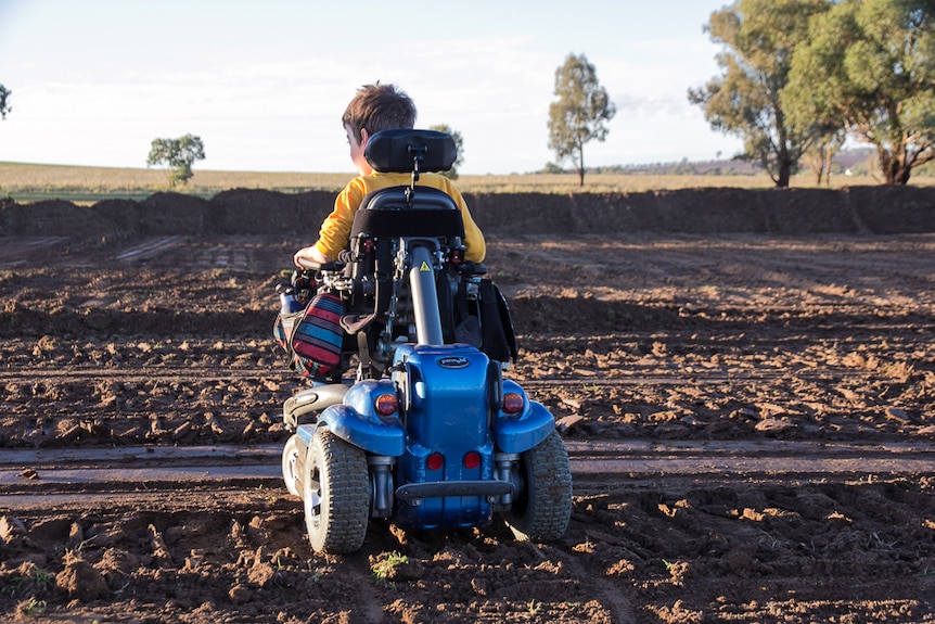 A boy in a blue power wheel chair on a flat block of cleared earth