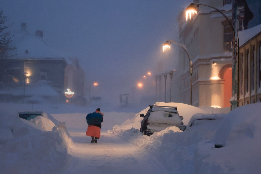 A person in winter clothing walks up a road covered in snow during a snowstorm in Kristiansand, Norway
