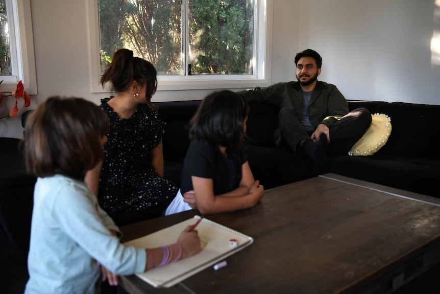 26-year-old man with dark hair, a trimmed beard, sits on couch chatting with three kids. Their heads are turned away from centre