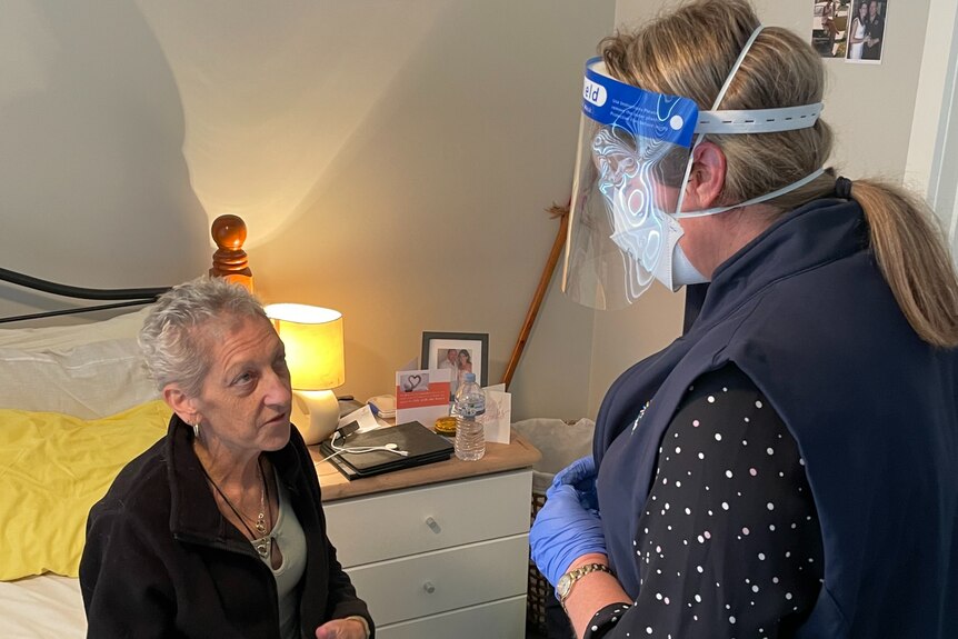 A woman with short gray hair wearing a black jacket sits on a bed and looks at a carer, who is wearing a face shield.