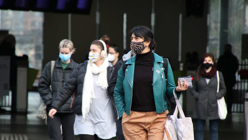 A woman in a green jacket, orange pants and patterned mask waits at a pedestrian crossing.