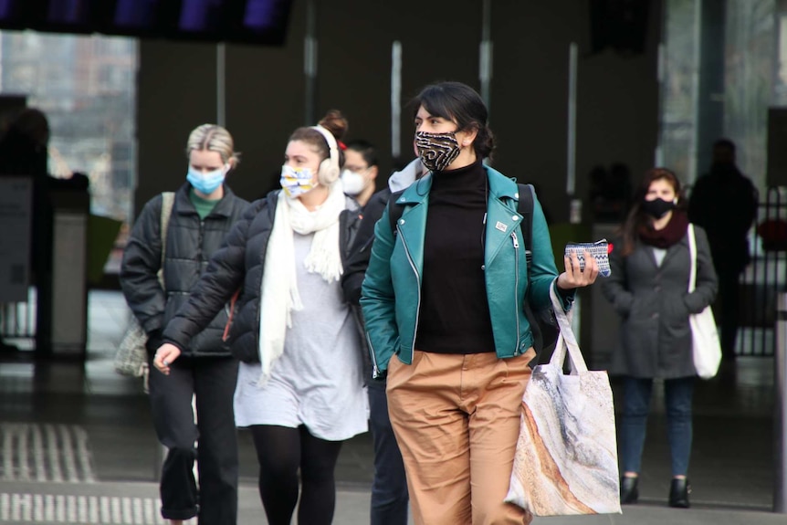 A woman in a green jacket, orange pants and patterned mask waits at a pedestrian crossing.