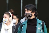 A woman in a green jacket, orange pants and patterned mask waits at a pedestrian crossing.
