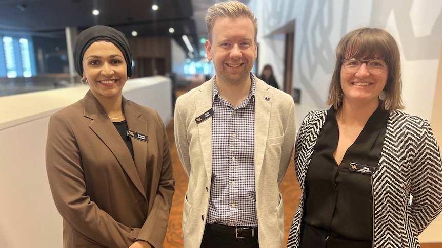 A male teacher stands between two female teachers in the corridor of a conference centre. They are smiling and formally dressed.