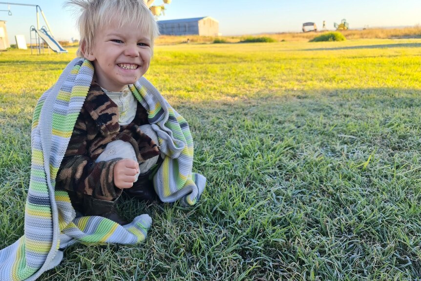 Toddler with a blanket wrapped around him looks at the frosty grass.