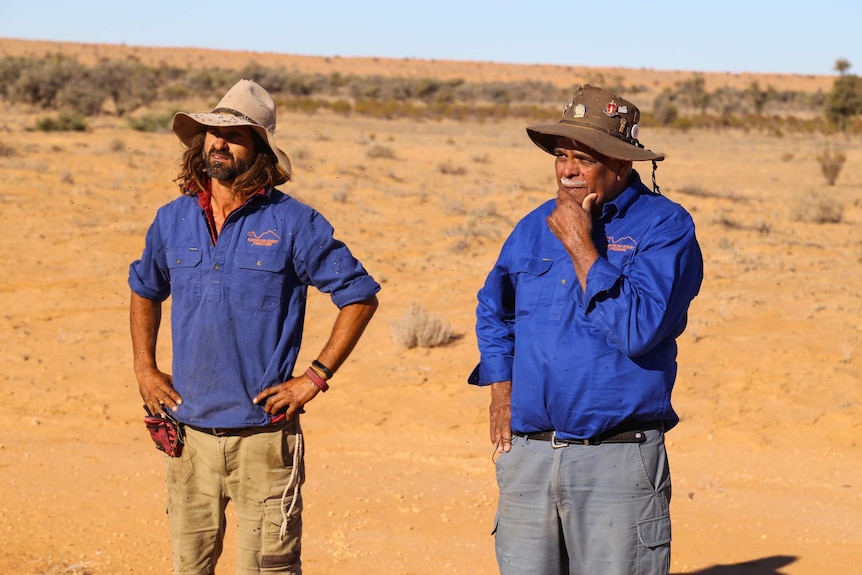 Two men wearing long-sleeved blue shirts and Akubra-style hats stand in the desert.