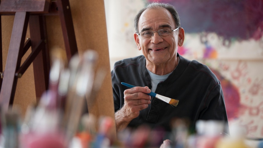 An older man holding a paintbrush smiles at the camera from behind his easel 