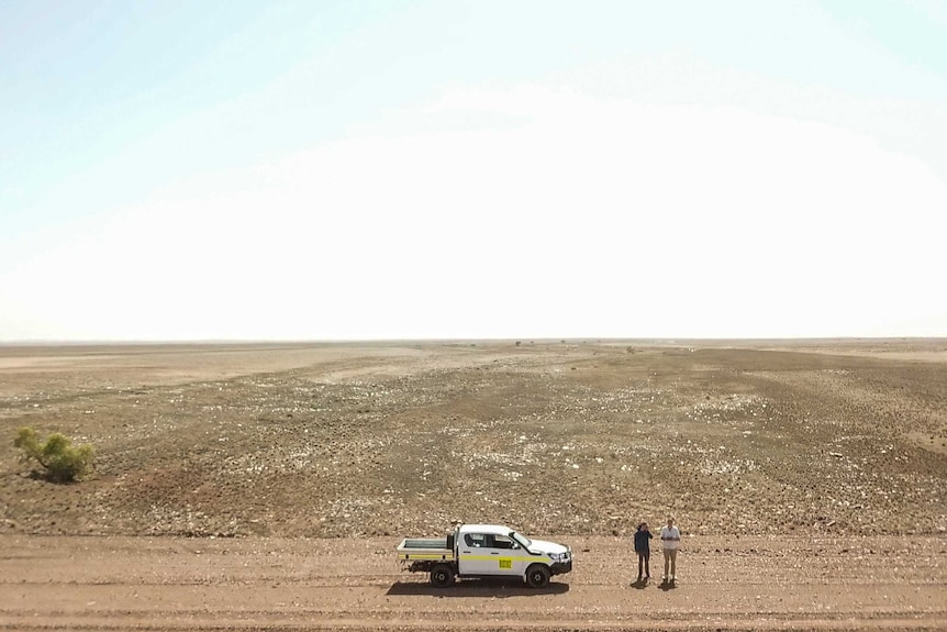 Two men stand by a white ute in the middle of a flat desolate desert plain.