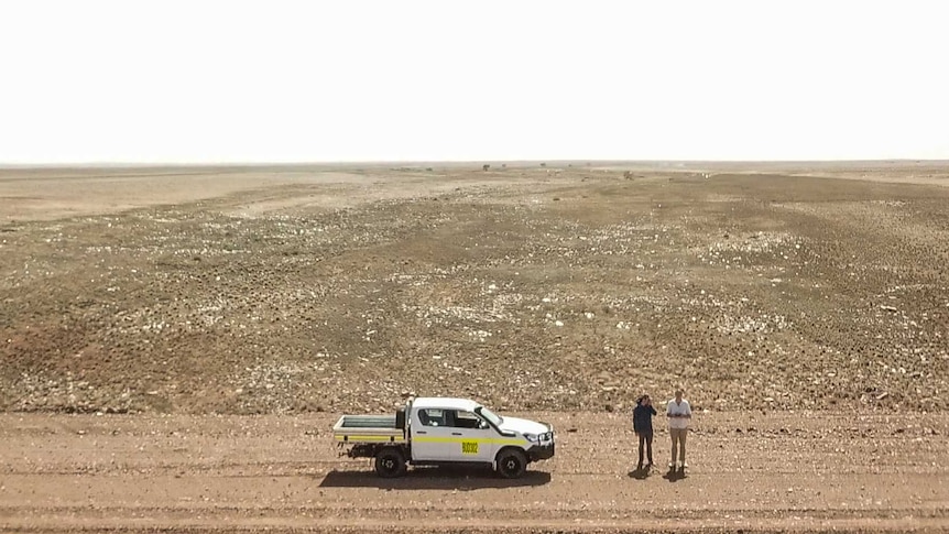Two men stand by a white ute in the middle of a flat desolate desert plain.