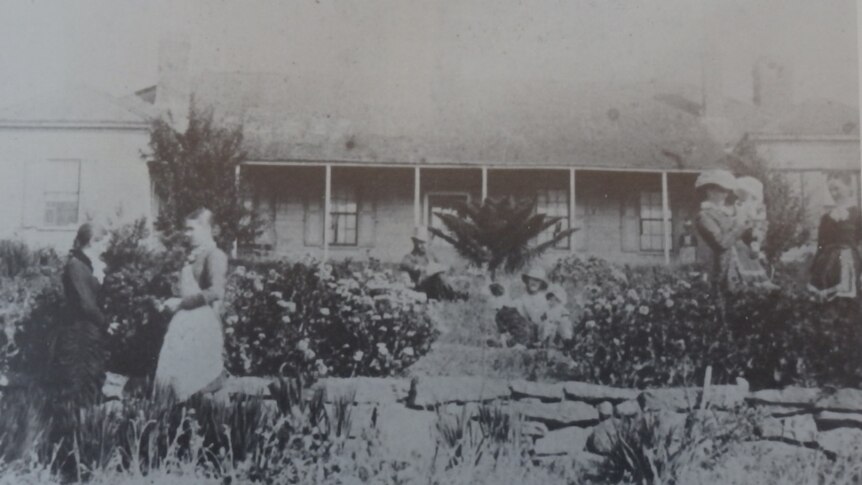 Grainy black and white photo of a family in late 1800s clothing standing outside a stone house