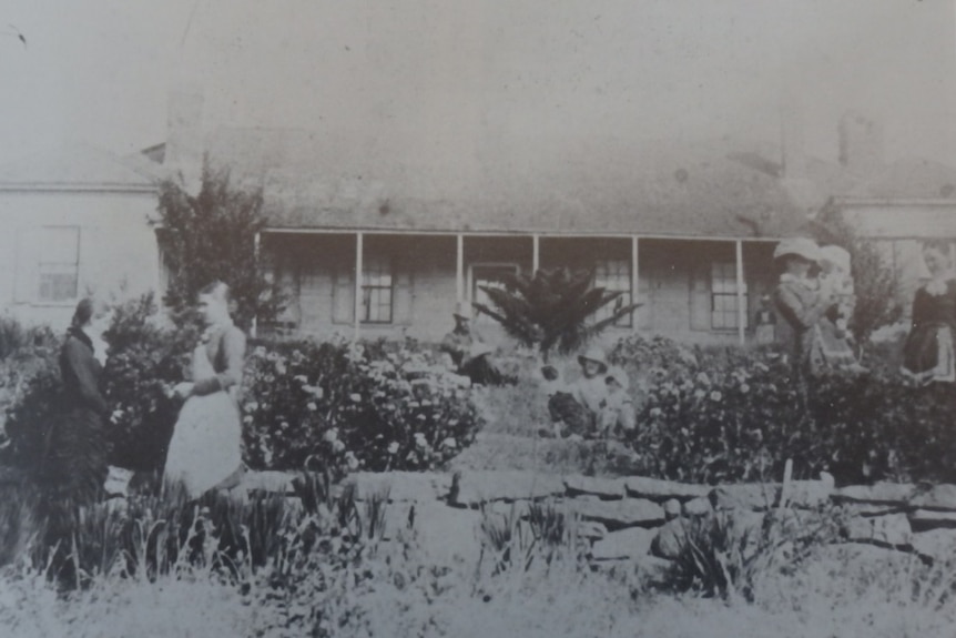 Grainy black and white photo of a family in late 1800s clothing standing outside a stone house