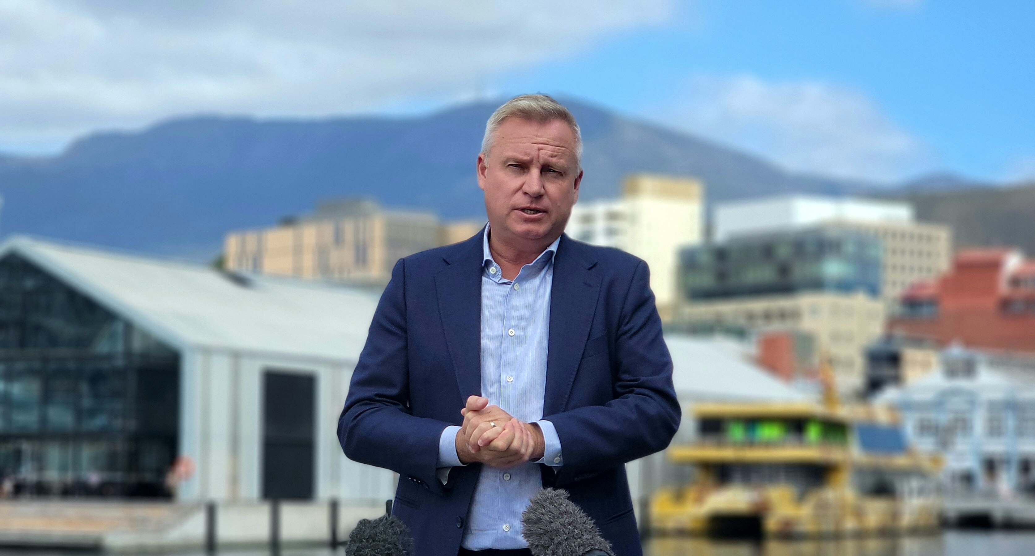 A middle aged man in a light blue shirt and dark blazer clasps his hands mid-speech. 