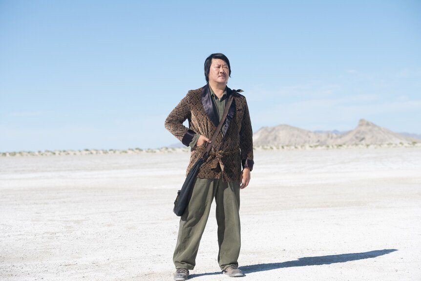 A 50 year old Chinese man in a suit jacket and green slacks with a bag slung across his shoulder stands in a desert