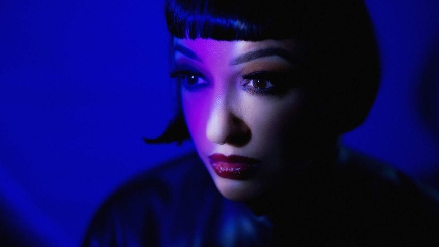 Carla Lippis stares away from the camera. She has a black bob haircut and red lipsitck. She's in front of a blue background.