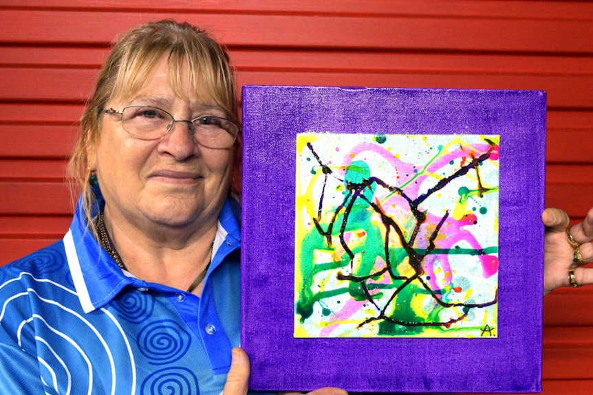 A woman standing in front of a red wall holds up a painting.