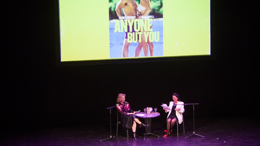 Myf Warhurst and Zan Rowe seated on stage in front of a poster of the movie 'Anyone But You'