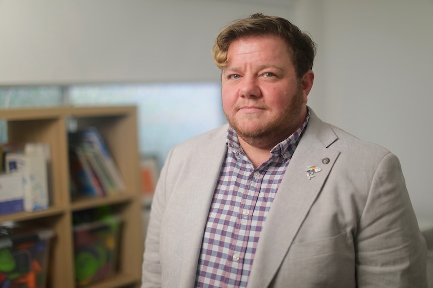 A person stands in an office, behind him are bookshelves. He is wearing a suit jacket and has a neutral expression.