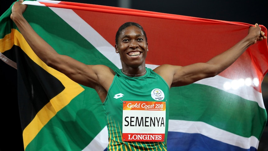 A black female athlete smiles as she holds up the South African flag behind her at a night-time athletics event.