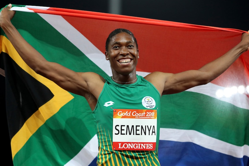 A black female athlete smiles as she holds up the South African flag behind her at a night-time athletics event.