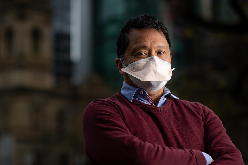 A man wearing a white duck-billed face mask and a maroon jumper over a blue shirt in front of 19th century buildings