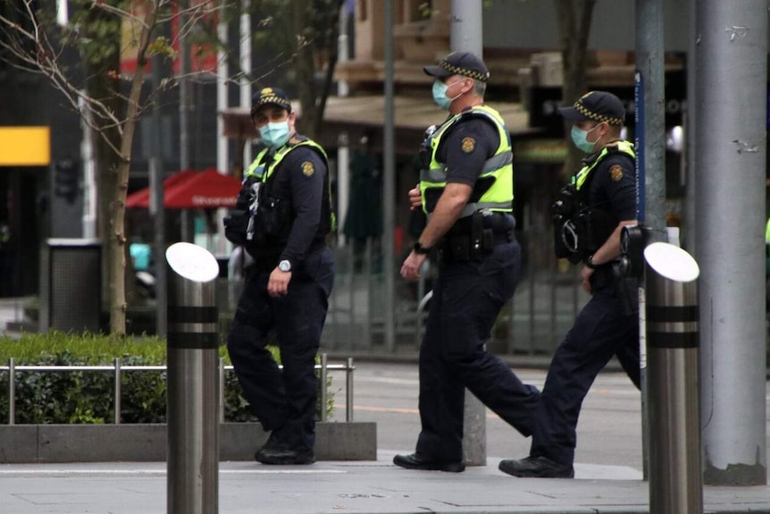 Three male police officers walk through the city, all wearing uniforms and masks.