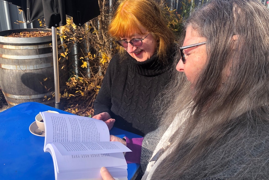 Woman with long graying hair reads a book at a coffee table in the garden with a smiling woman with orange hair wearing a black sweater.