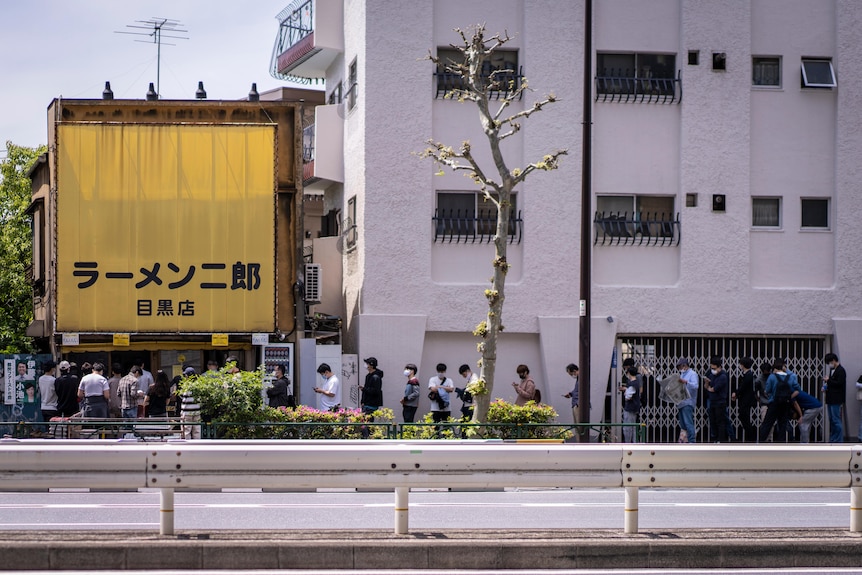 A row of people outside a building with Japanese writing on the side 