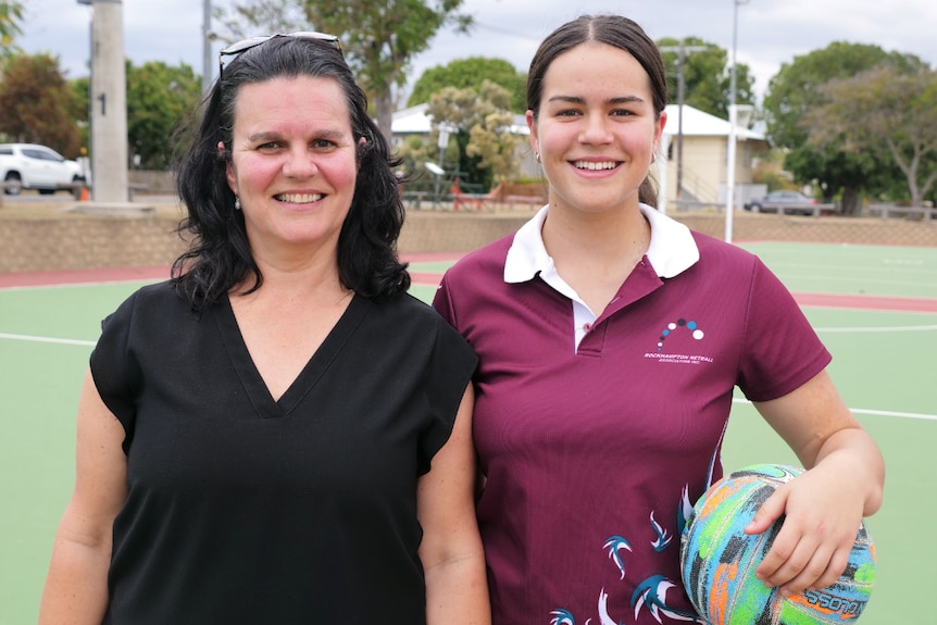 A mum and daughter stand together on the netball court smiling.