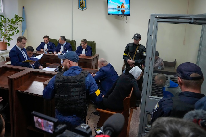 a small courtroom. men is suits sit around the room. in a transparent box, the russian sits, leaning forward