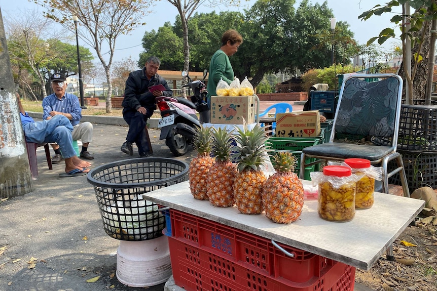 Some fresh and bottled pineapples sit on a table with old people sitting around.