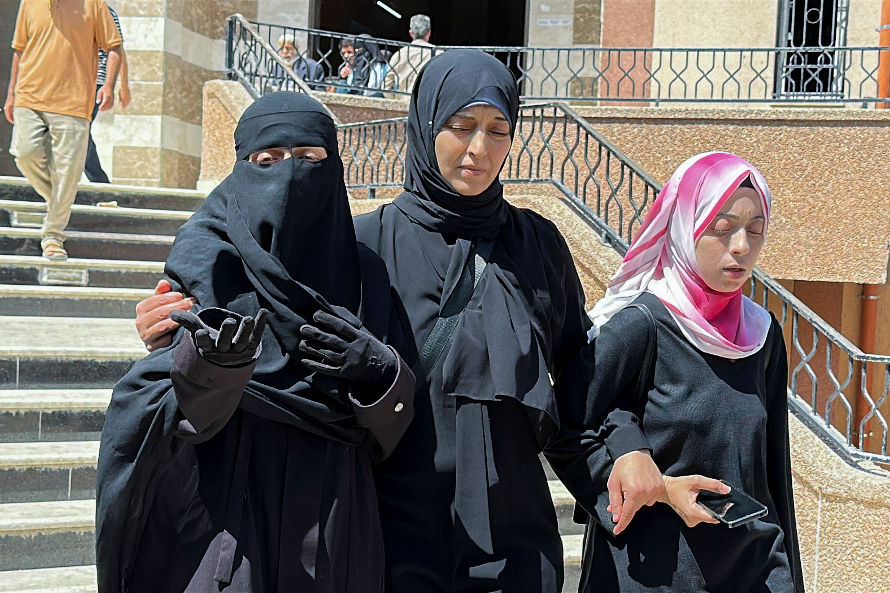 Three women wearing head dresses looking distressed on a staircase, with a woman on the far left gesturing with her hands