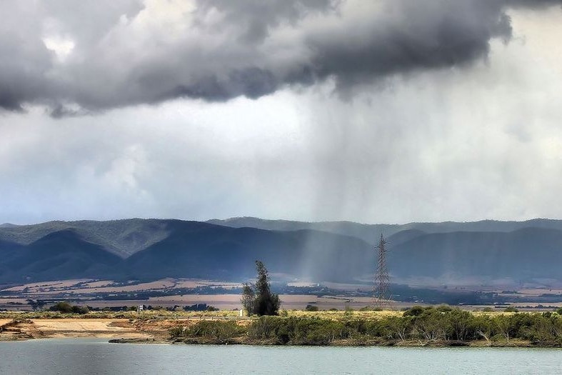 A heavby rain cloud sits above a mountain range behind farmland, with a bay of water in the foreground.