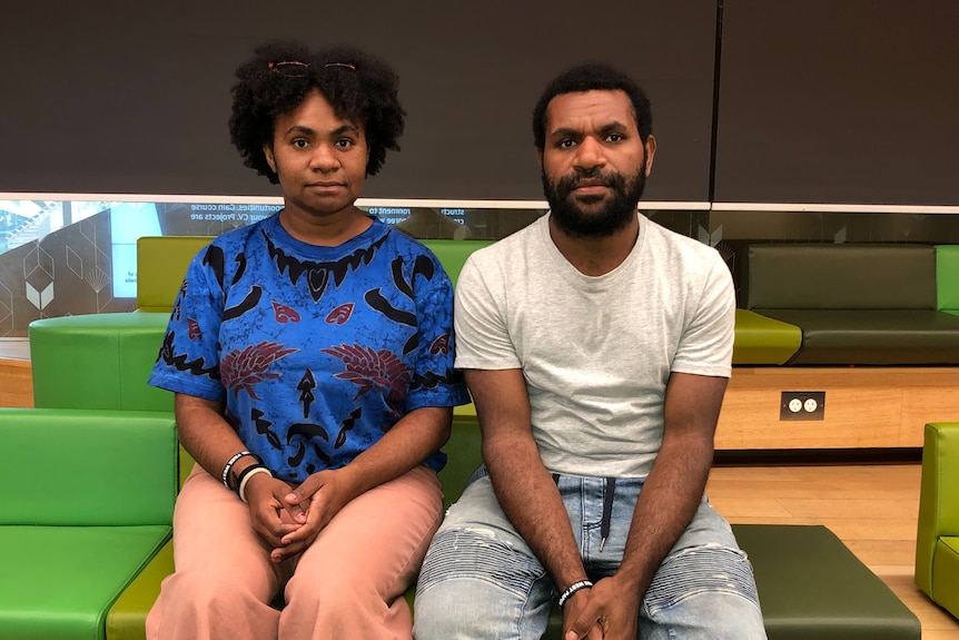 A West Papuan man and a woman sit on a low couch looking at the camera unsmiling.