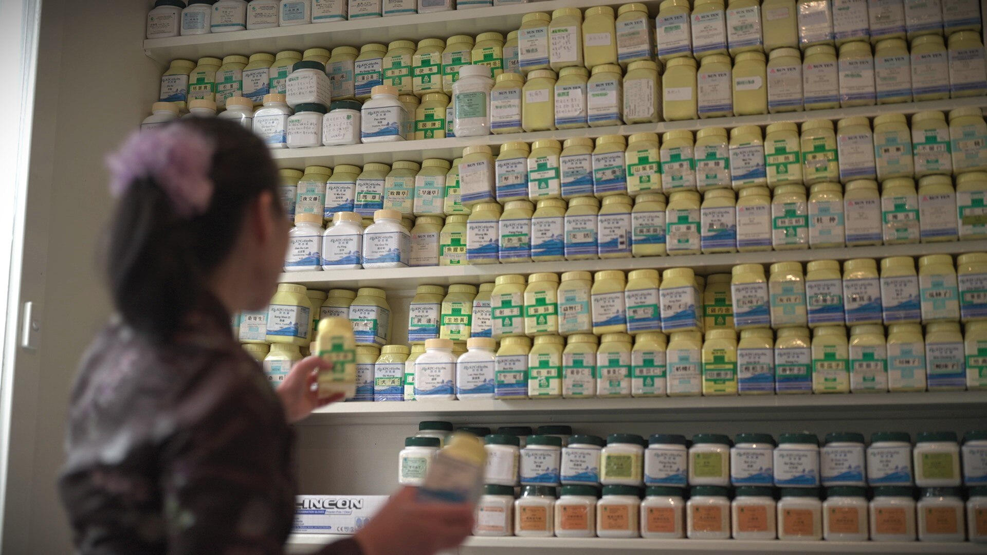 An elderly Chinese woman wearing a purple scrunchy in her hair browses a shelf of Chinese medicine.