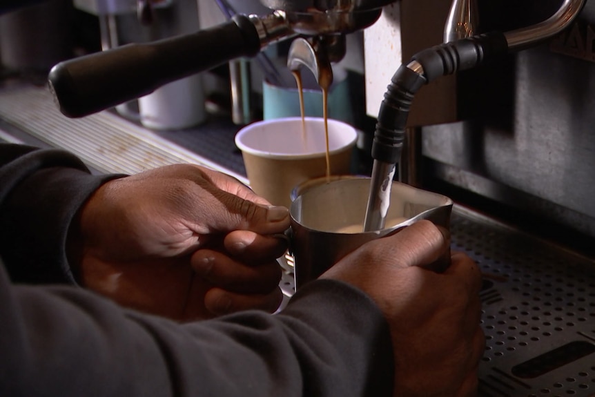 A pair of hands steaming a stainless steel jug of milk at an espresso machine.