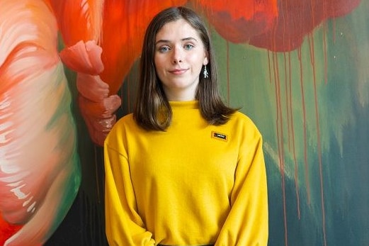Bri Hines stands in front of a wall with a colourful painting on it.