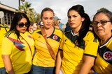 A group of Hispanic women in yellow shirts stand together looking serious.