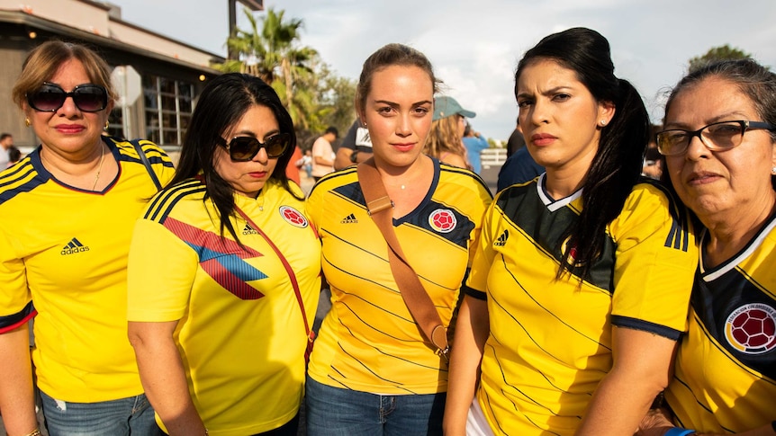 A group of Hispanic women in yellow shirts stand together looking serious.