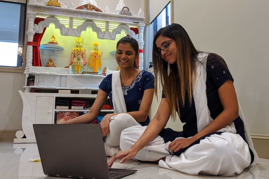 Hemangini Patel and another young woman sitting with a laptop, with an in-home  Hindu altar in the background.